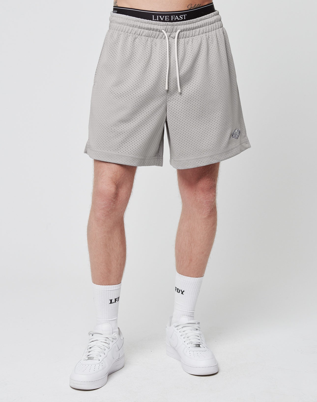 A person stands against a plain white background wearing LIVE FAST DIE YOUNG Basic Mesh Shorts (gray, logo patch, regular fit), white LIVE FAST socks, white sneakers, and black LIVE FAST underwear. Upper body not visible.