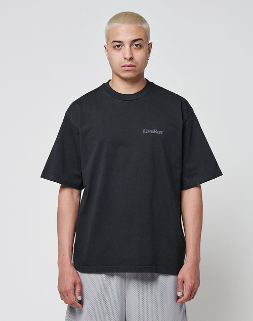 A person with short bleached blond hair wears a loose black LIVE FAST DIE YOUNG Basic 280 Tee with backprint and gray mesh shorts in an oversize fit, standing against a plain white background.