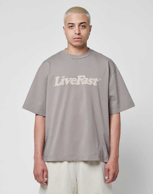 A person with short, bleached blonde hair wears the LIVE FAST DIE YOUNG Basic Tee in taupe, Oversize Fit aus Baumwolle mit Vintage-Waschung and LiveFast print, paired with light mesh shorts, against a plain white background.
