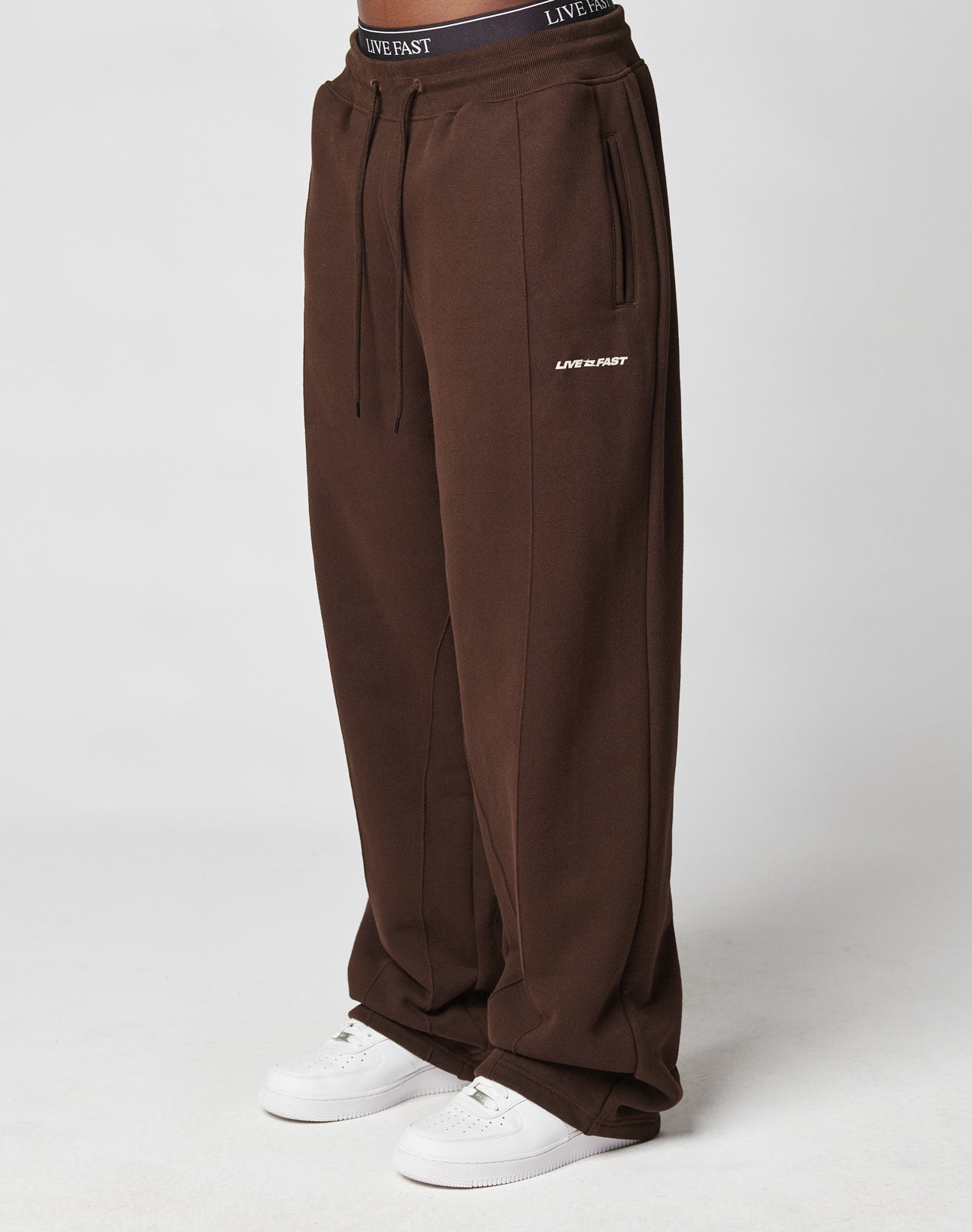 Someone wearing LIVE FAST DIE YOUNG LFDY Pleated Jogger wide fit brown sweatpants with LIVE FAST text on the left thigh, elastic waistband, and white sneakers, stands against a plain light background.