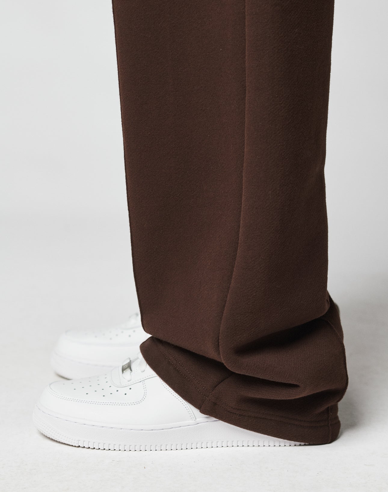 A close-up side view of a person wearing the LFDY Pleated Jogger by LIVE FAST DIE YOUNG, featuring a wide fit and Elastik-Bund in brown, draped over white perforated sneakers against a plain light backdrop.
