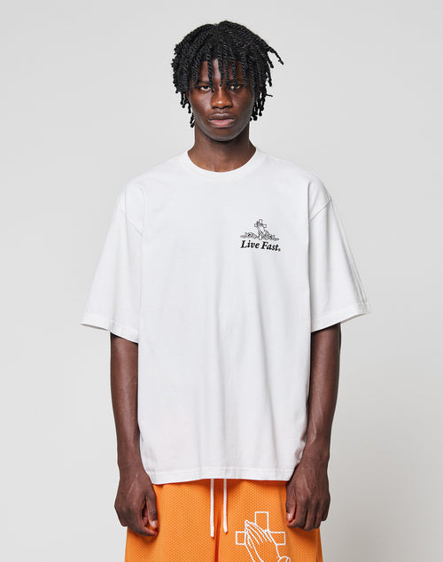 A person with medium-length braided hair wears the LFDY Praying Tee by LIVE FAST DIE YOUNG, featuring “Live Fast.” front and back prints, paired with orange basketball shorts with a white cross design, against a light gray background.