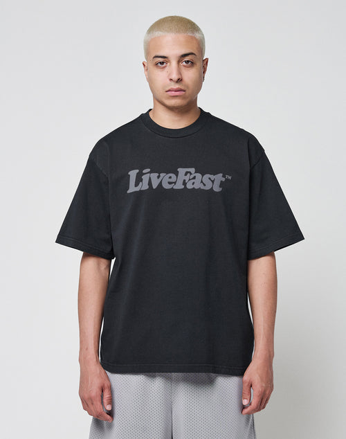 A person with short blond hair wears a loose black Basic Tee from LIVE FAST DIE YOUNG in an oversize fit, featuring LiveFast printed in gray on the front, standing against a plain light background.