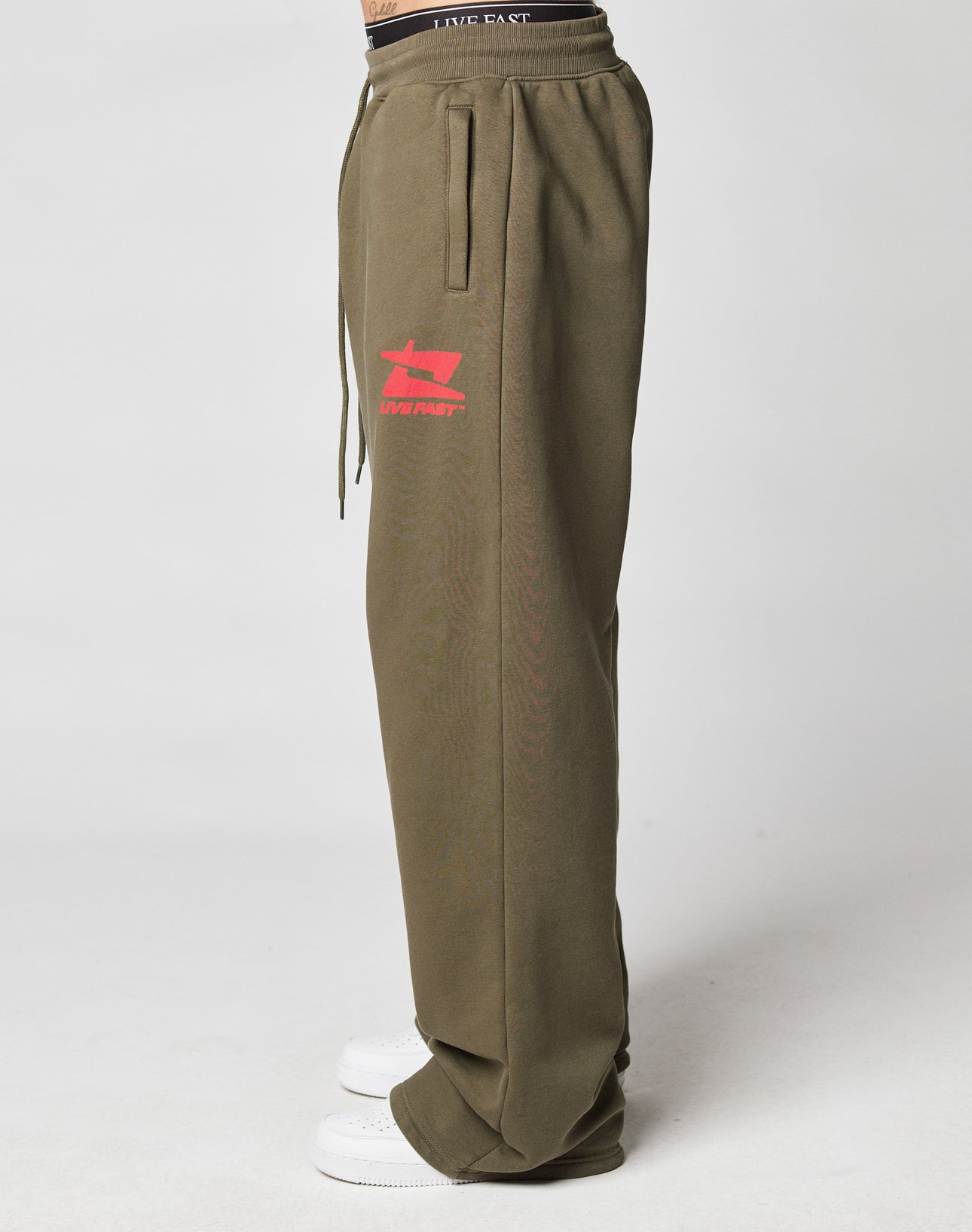 Someone stands sideways against a plain light background, wearing the olive green LIVE FAST DIE YOUNG Live Fast Sweatsuit Jogger with a Breiter Elastik-Bund and bold red LIVE FAST graphic, paired with white sneakers.