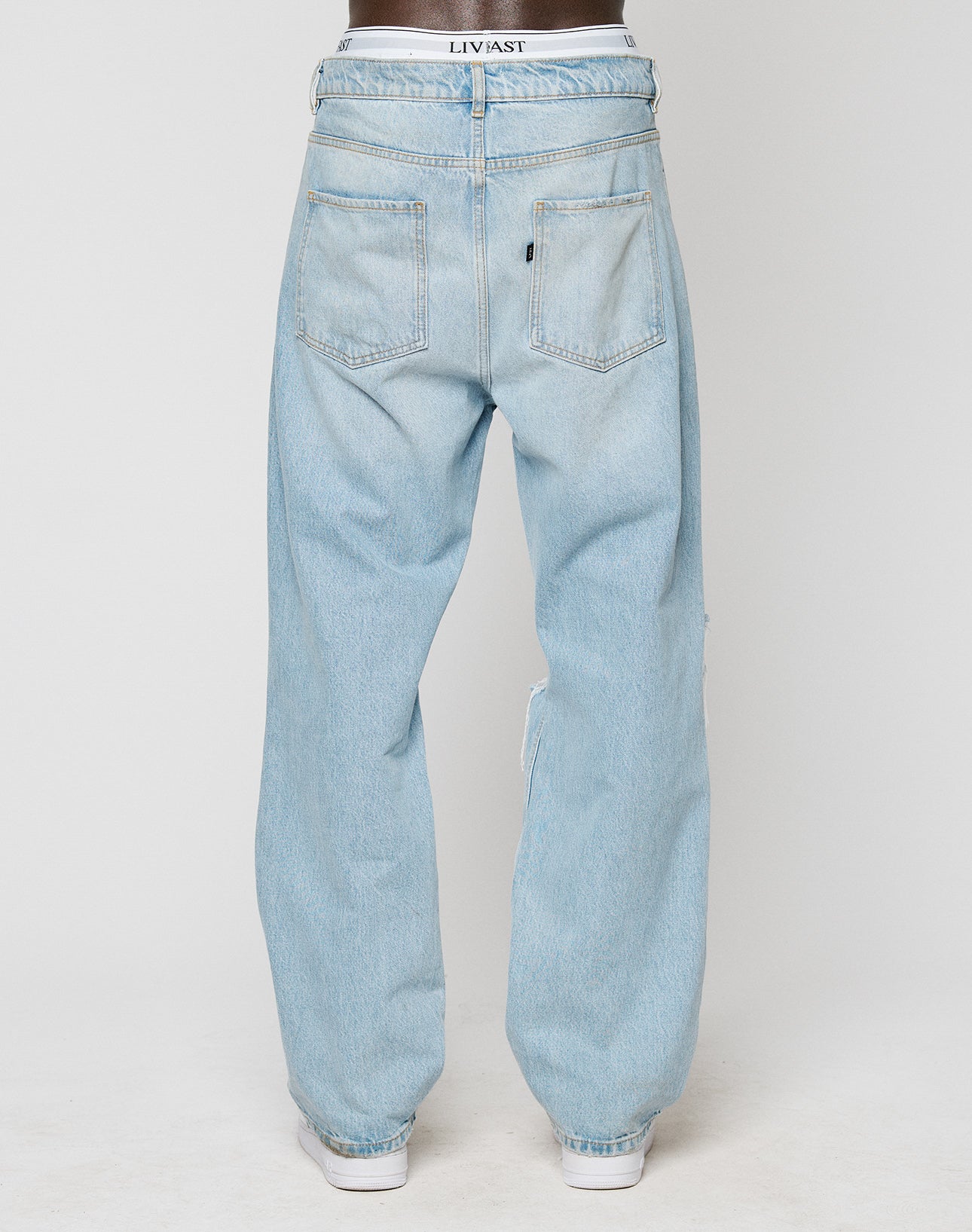 A person is seen from the back wearing LFDY Distressed Baggy Denim jeans by LIVE FAST DIE YOUNG. The loose, light blue jeans have two large back pockets, visible seams, white shoes, and partially visible LEVIS waistband underwear.