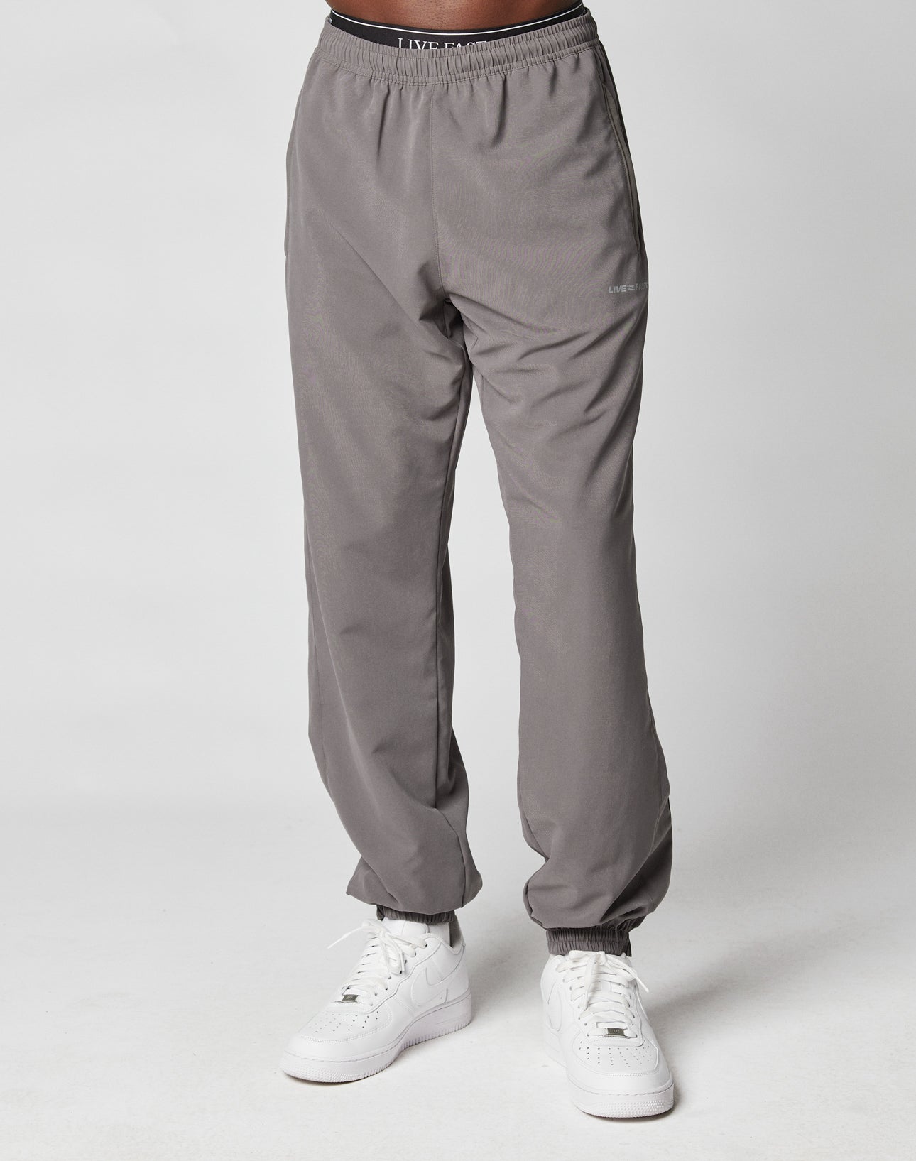 A person wears LIVE FAST DIE YOUNG LFDY Trackpants with Elastikbund and white sneakers, standing against a plain white background. Only the lower half is visible, highlighting the pants casual Regular Fit.