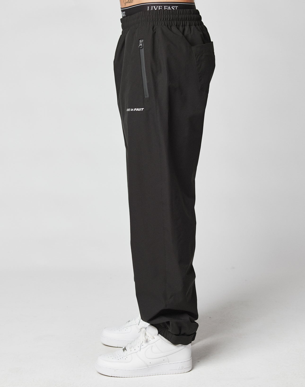 A person stands sideways in Basic Shell Pants by LIVE FAST DIE YOUNG, featuring reflektierender Druck, a zipper pocket, and LIVE FAST on the left thigh. The joggers have an Elastikbund mit Kordelzug and are styled with white sneakers against a plain white background.