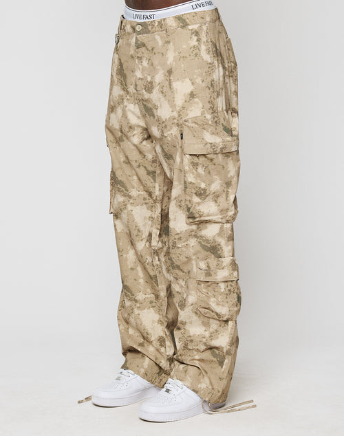 A person wears LFDY Cargo by LIVE FAST DIE YOUNG: loose-fitting tan camo cargo pants with Cargo Taschen, Wide Fit, and a white LIVE FAST waistband. Theyre paired with white sneakers. Made from 100% Baumwolle; backdrop is plain light gray.