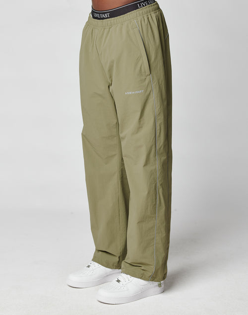 Someone wearing loose-fitting, olive green Live Fast Trackpants from LIVE FAST DIE YOUNG, featuring branding on the waistband and left pocket, pairs them with white sneakers and stands against a plain white background.