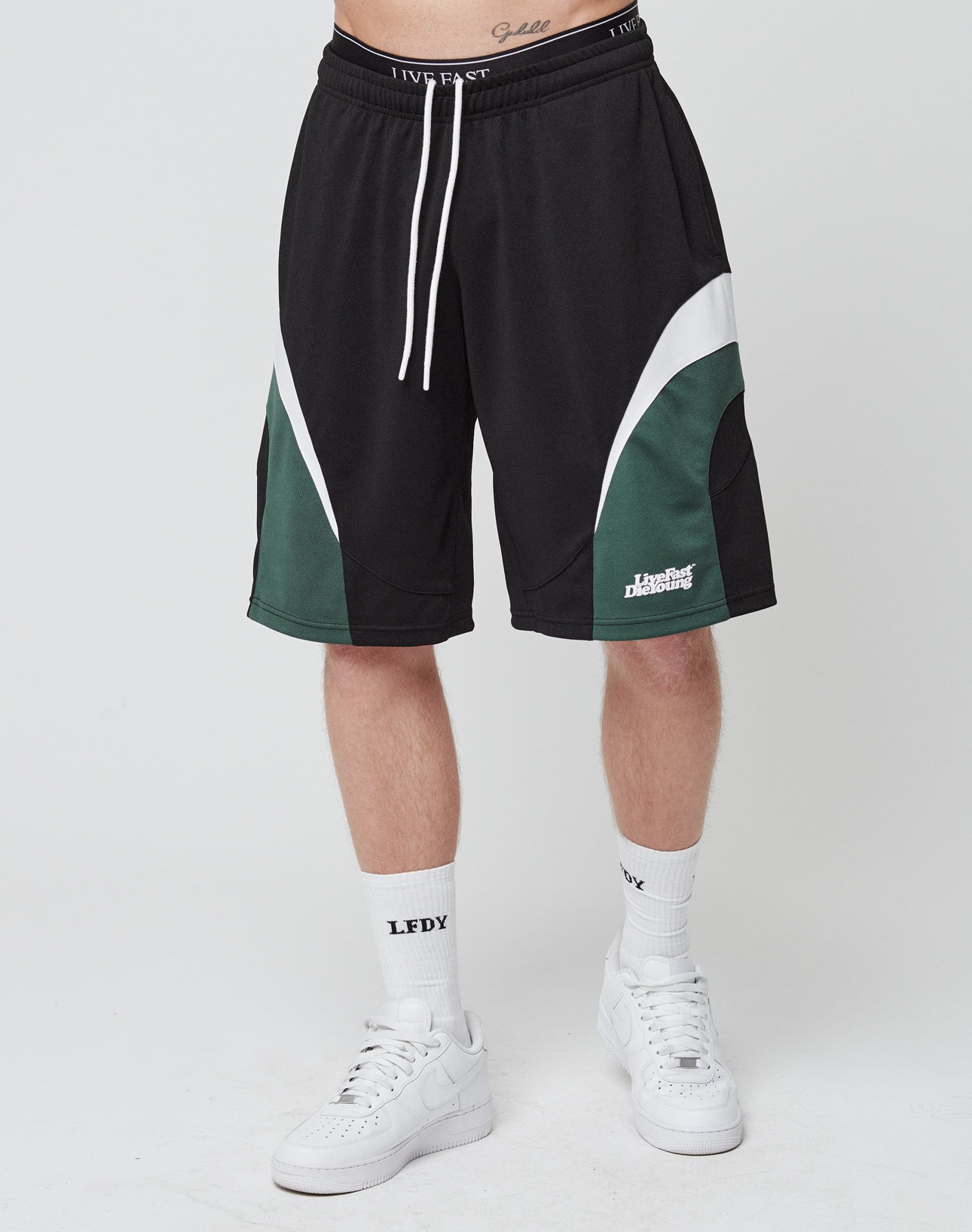 A person modeling LIVE FAST DIE YOUNG Basic Trackshorts in black, white, and green with elastic waistband, paired with LFDY Logo-Print socks and white sneakers, stands against a plain white background. Only the lower body is visible.