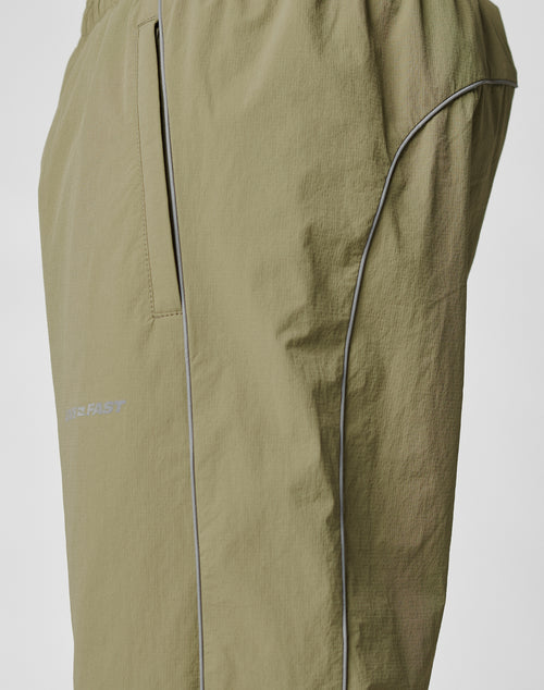 Close-up of the Live Fast Trackpants by LIVE FAST DIE YOUNG in light olive green, showcasing a zippered side pocket, reflective logo print, piping along the seams, and an elastic drawcord waist against a plain background.