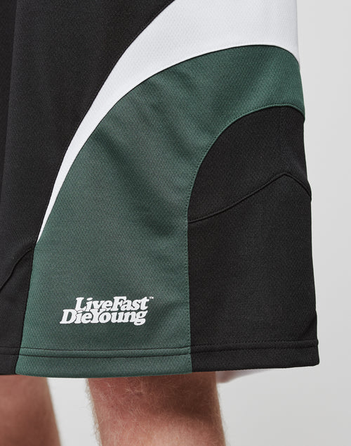 Close-up of black Basic Trackshorts by LIVE FAST DIE YOUNG, featuring green and white curved panels, a logo print, and Live Fast Die Young text in white on the lower left near the elastic waistband; part of a leg is visible.