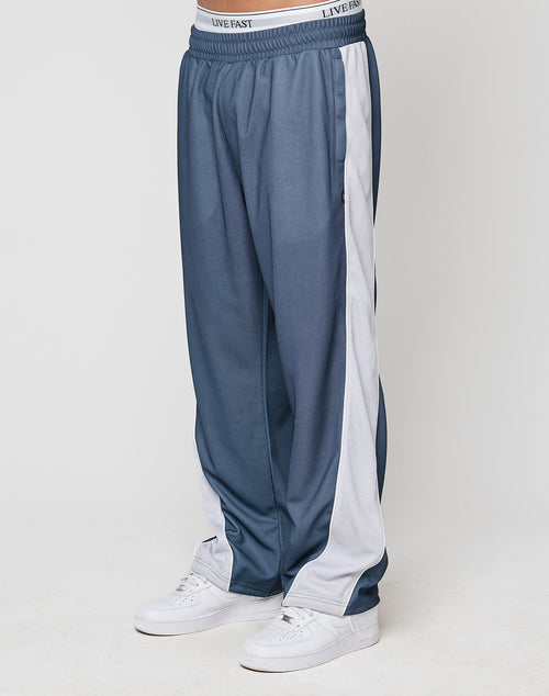 A person stands against a plain white background wearing LFDY DY Trackpants by LIVE FAST DIE YOUNG—wide fit blue pants with a white elastic waistband reading LIVE FAST, white side stripes, and paired with white sneakers.
