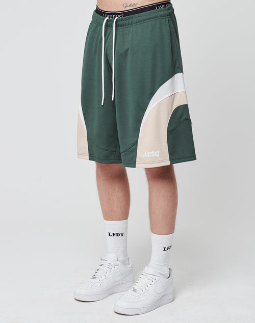 A person wears LIVE FAST DIE YOUNG Basic Trackshorts in green and beige (100% polyester), white LFDY socks, and white sneakers. Only their lower half is visible against a plain white background.