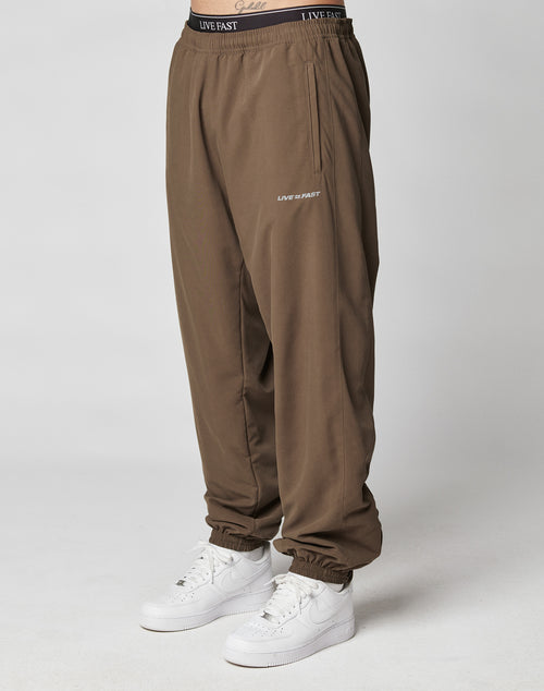 A person wearing LFDY Trackpants by LIVE FAST DIE YOUNG, featuring an elastic waistband and LIVE FAST reflective print on the band and thigh, paired with white Nike sneakers, stands before a plain light background.