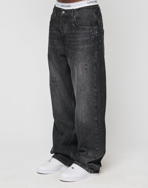 Someone wears the LFDY Baggy Denim by LIVE FAST DIE YOUNG—black jeans with wide straight legs, light fades, unique color splatters, subtle distressed details—paired with white sneakers and a visible LIVE FAST waistband.