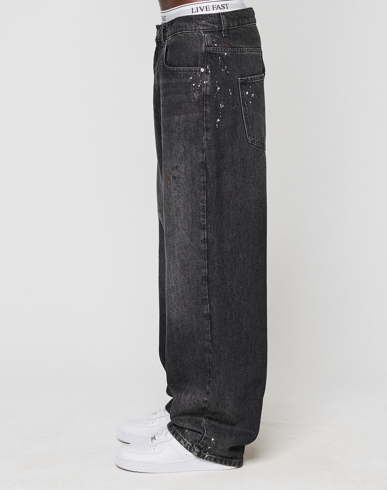 A person in LIVE FAST DIE YOUNG’s LFDY Baggy Denim—baggy fit, dark gray with individuelle Farbspritzer—paired with white sneakers and a LIVE FAST waistband, seen from the side against a plain background.