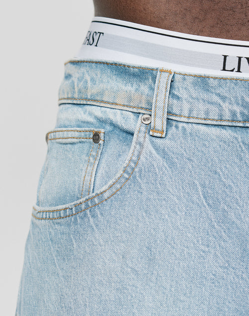 Close-up of a person wearing LFDY Baggy Denim jeans from LIVE FAST DIE YOUNG, with a focus on the pocket and waistband area where white underwear with black stripes and partial black text is visible.