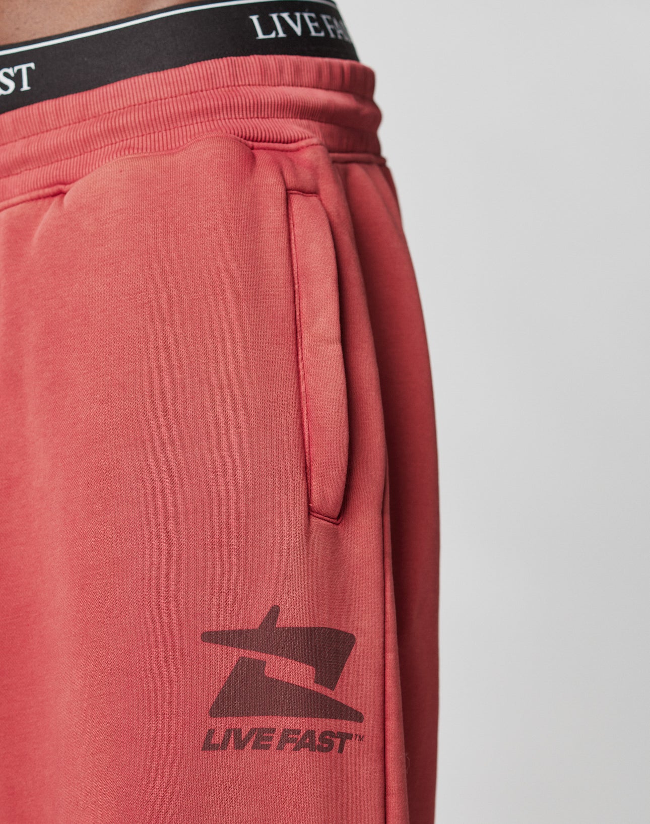 Close-up of a person in LIVE FAST DIE YOUNG Live Fast Sweatsuit Jogger—red, vintage-wash joggers with deep pockets and a black LIVE FAST logo near the hip. Black underwear waistband with LIVE FAST text peeks above the elastic waist.