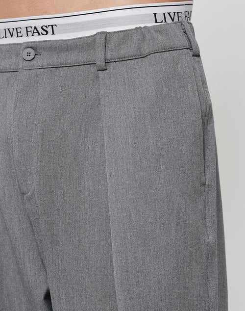 Close-up of a person in Wide Casual Pants by LIVE FAST DIE YOUNG, featuring a straight leg and subtle pleats. The elastic waistband of white underwear with LIVE FAST in black shows above the pants.