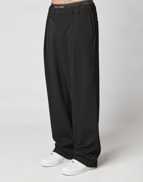 A person wears LIVE FAST DIE YOUNG Wide Casual Pants—loose, wide-fit black trousers with side pockets and an elastic waistband featuring LIVE FAST—paired with white sneakers; only the lower half is shown against a light background.