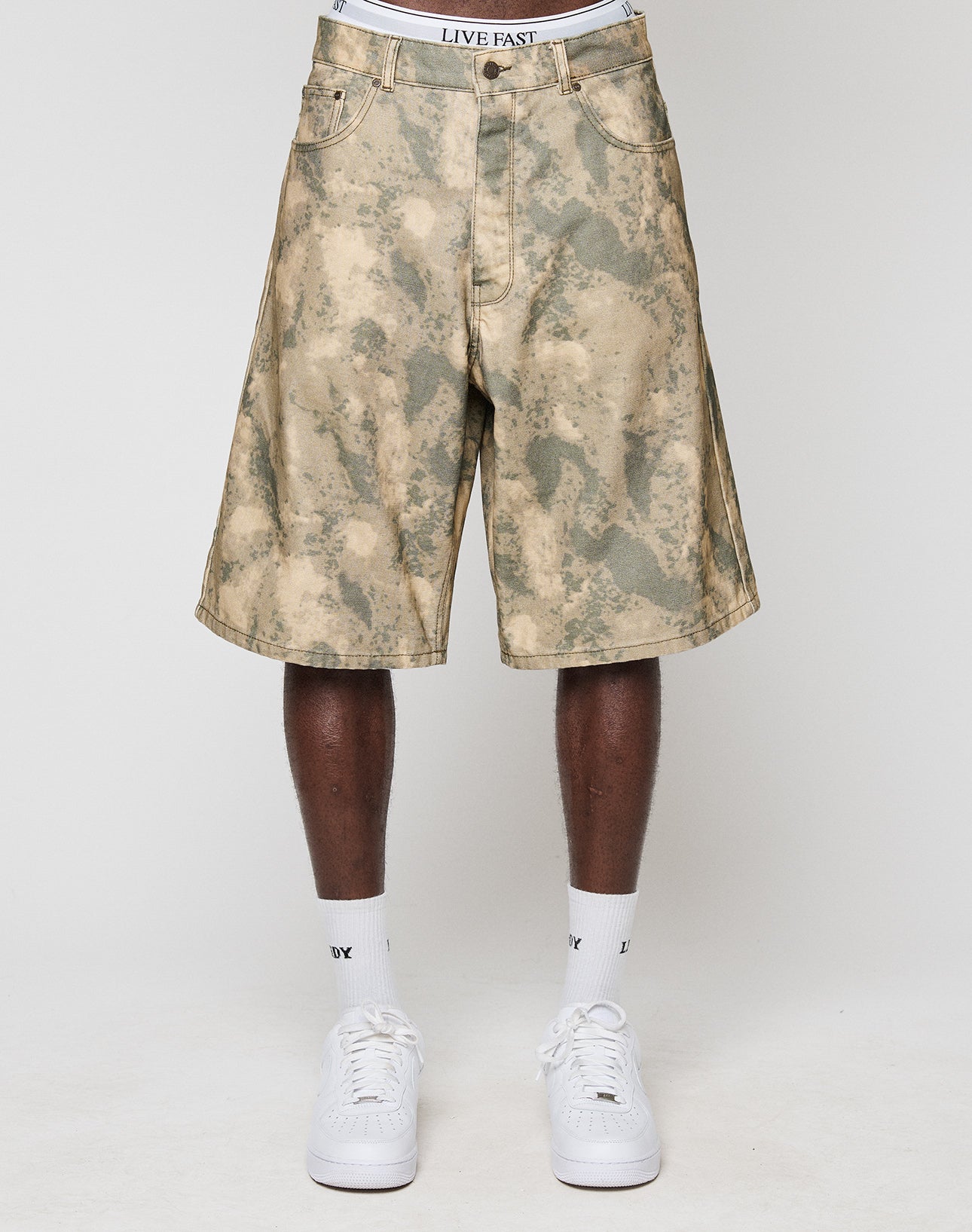 A person wears tan and green camouflage Canvas Shorts by LIVE FAST DIE YOUNG with a baggy fit and straight leg, white socks, and white sneakers on a plain white background. The LIVE FAST waistband of their underwear shows above the shorts.
