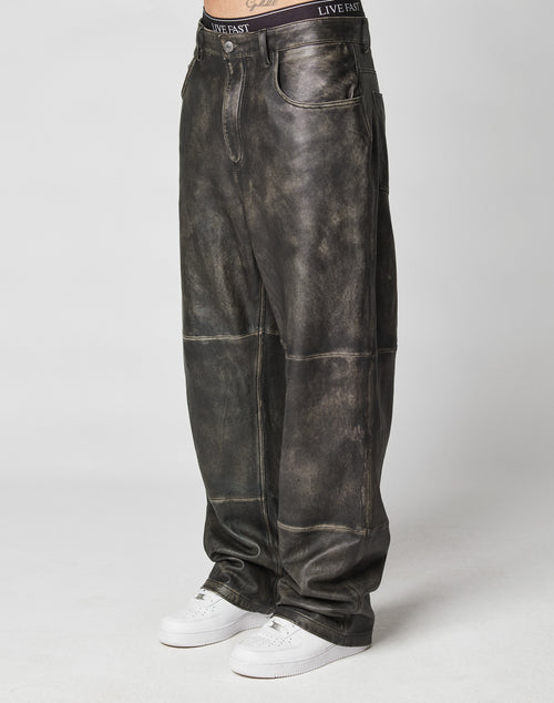 A person showcases the lower body in LIVE FAST DIE YOUNG Baggy Leather Pants—dark distressed, visible seams, and vintage style—paired with white sneakers against a plain background.
