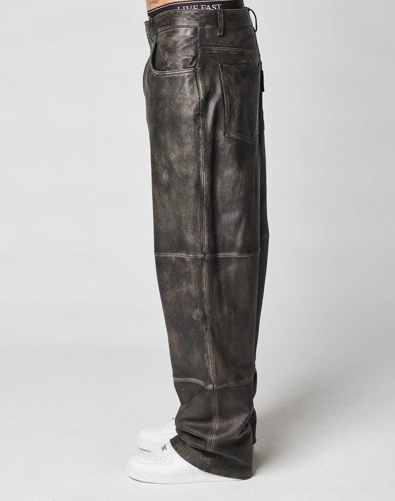 Side view of a person in baggy fit, distressed black Baggy Leather Pants by LIVE FAST DIE YOUNG with a flag label, white sneakers, and a black waistband that features “LIVE FAST,” standing against a plain light background.