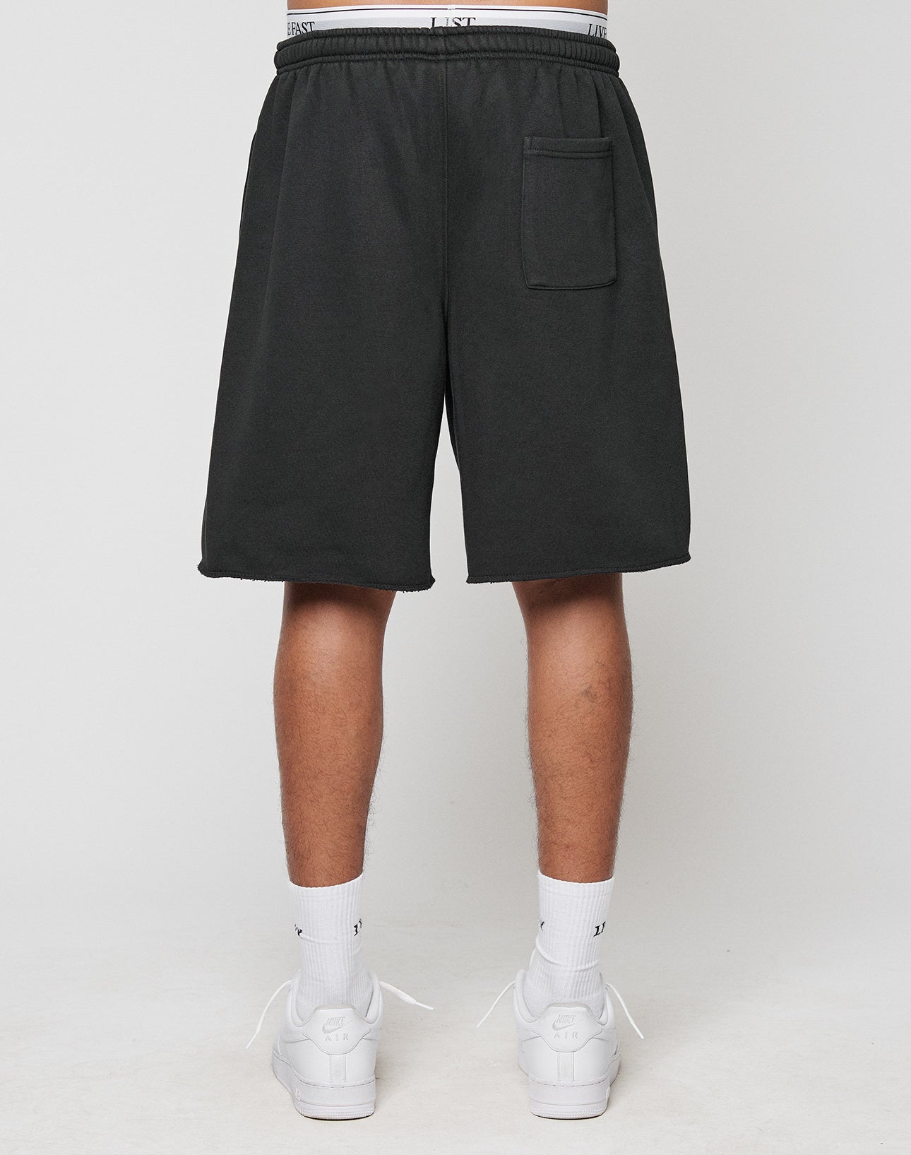 A person stands facing away, wearing black LFDY Praying Shorts by LIVE FAST DIE YOUNG with side pockets and a back pocket, paired with white crew socks and sneakers against a plain light background.