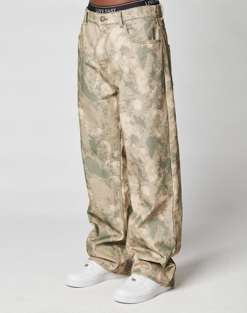 A person models LIVE FAST DIE YOUNGs Live Fast Canvas Pants—baggy fit, 100% cotton, light tan and green camo print with straight legs—paired with white sneakers and black waistband underwear reading LIVE FAST, set against a plain white background.