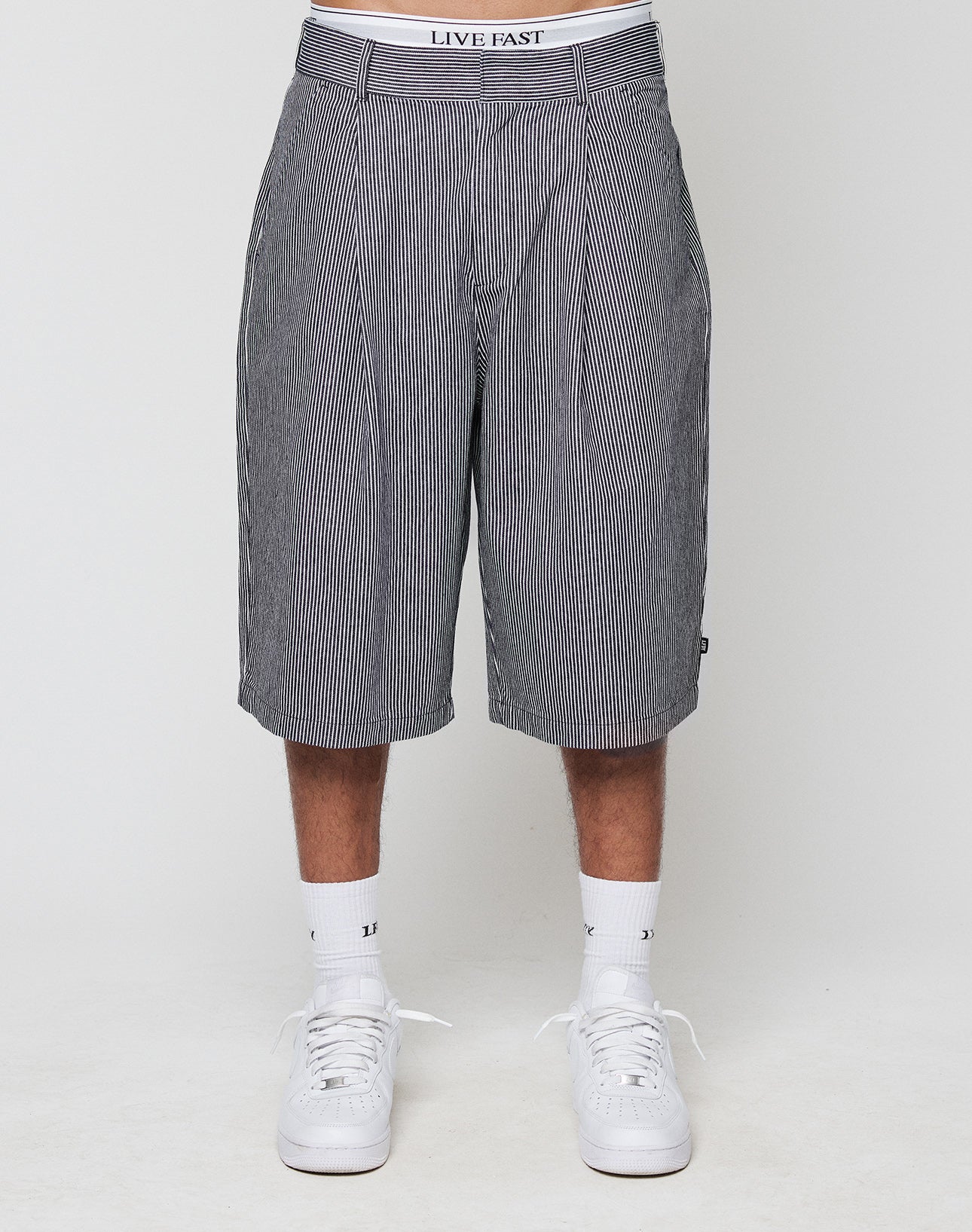 A person wears black and white pinstriped LFDY Twill Shorts by LIVE FAST DIE YOUNG, paired with white sneakers, socks, and LIVE FAST waistband underwear. Photo is cropped from mid-torso down.