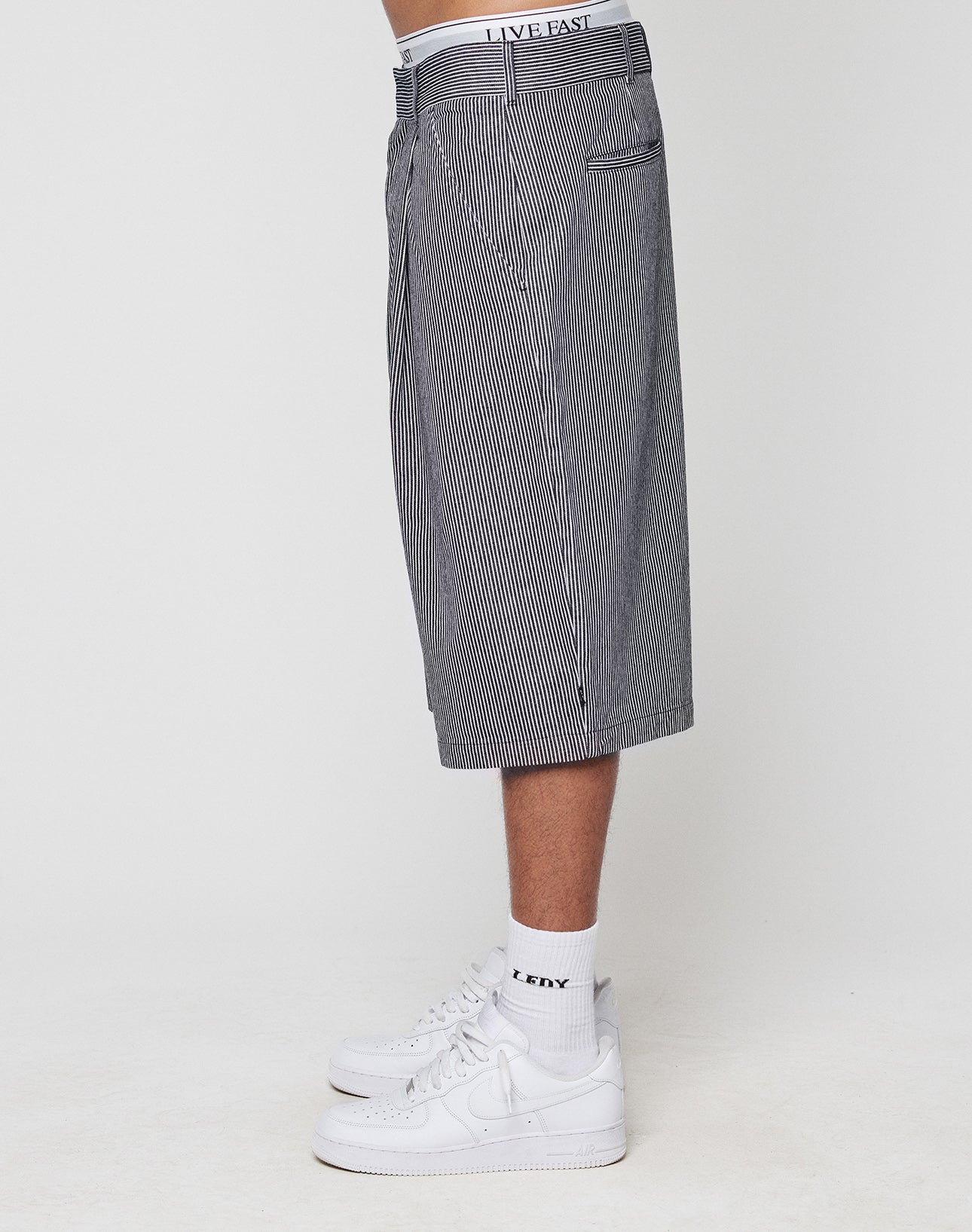 A person stands in profile against a plain white background wearing LFDY Twill Shorts by LIVE FAST DIE YOUNG with an oversize fit, wide straight leg, LIVE FAST waistband text, white crew socks with EXXY text, and white sneakers.