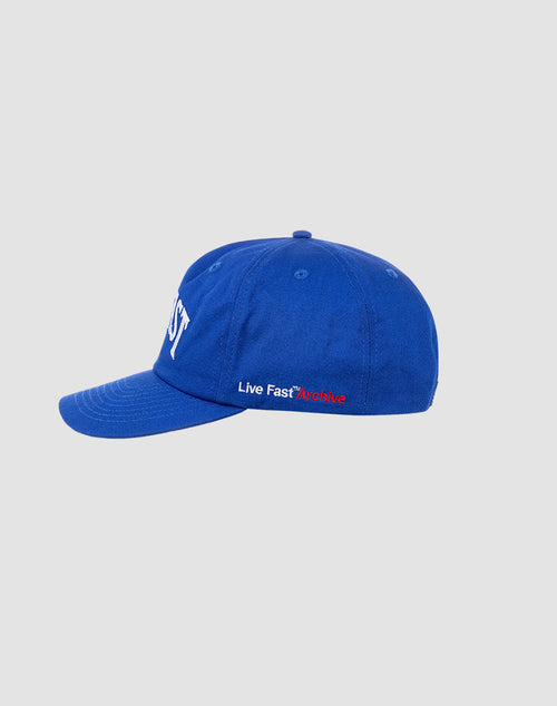 The LIVE FAST DIE YOUNG Live Fast Unstructured Cap is blue with white front text, a Flag-Label detail, and “Live Fast*Archive” embroidered in white and red on the side. It features a Mit Snapverschluss and is shown on a plain light gray background.