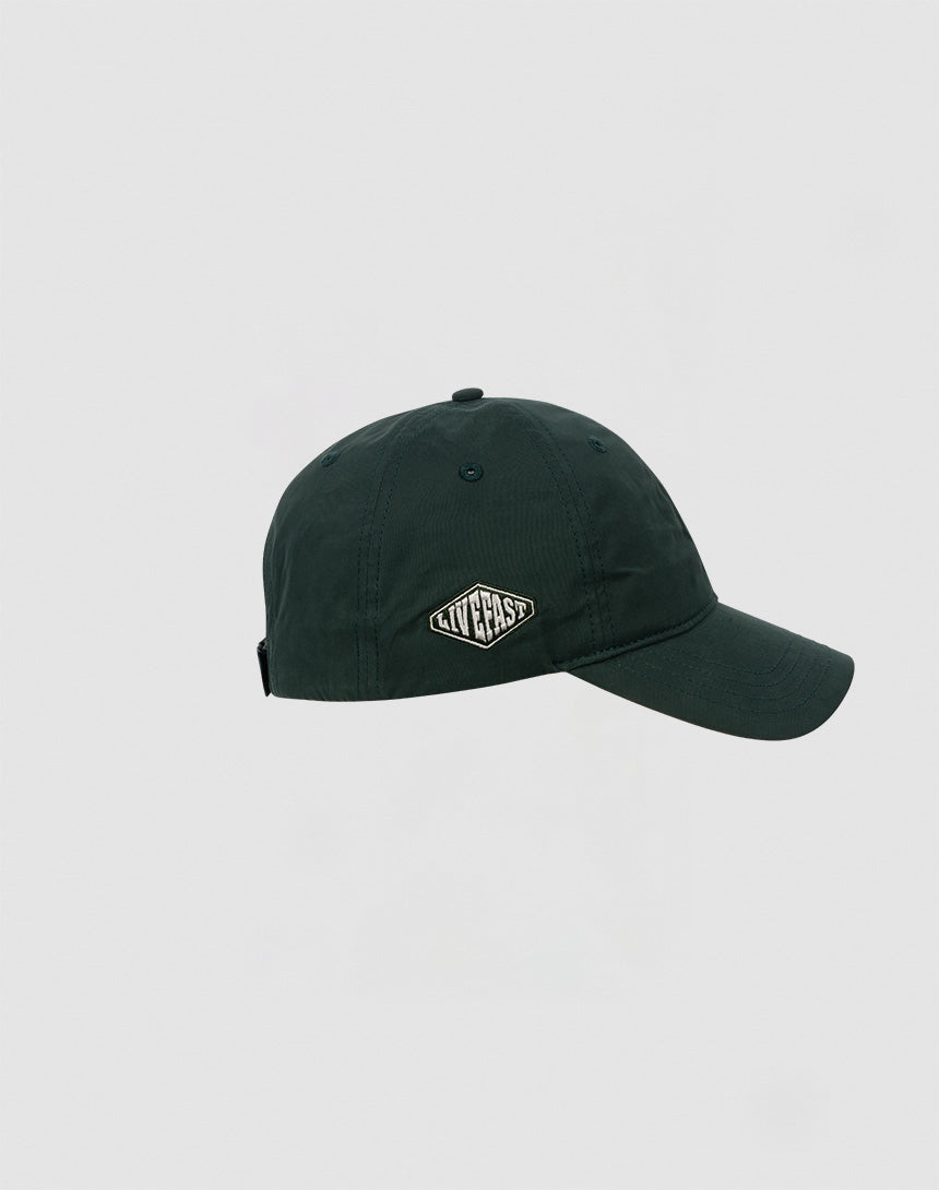 A dark green Basic Cap by LIVE FAST DIE YOUNG made of polyester with a curved brim and a small oval patch on the front displaying the brand name in white letters. Shown against a plain light gray background.