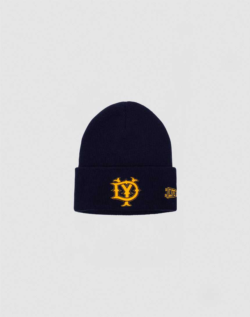 The DY Beanie by LIVE FAST DIE YOUNG is a navy blue polyacryl beanie with a folded brim, featuring a yellow Logo-Stick Y crest on the front and a small yellow text patch on the side, set against a plain light gray background.