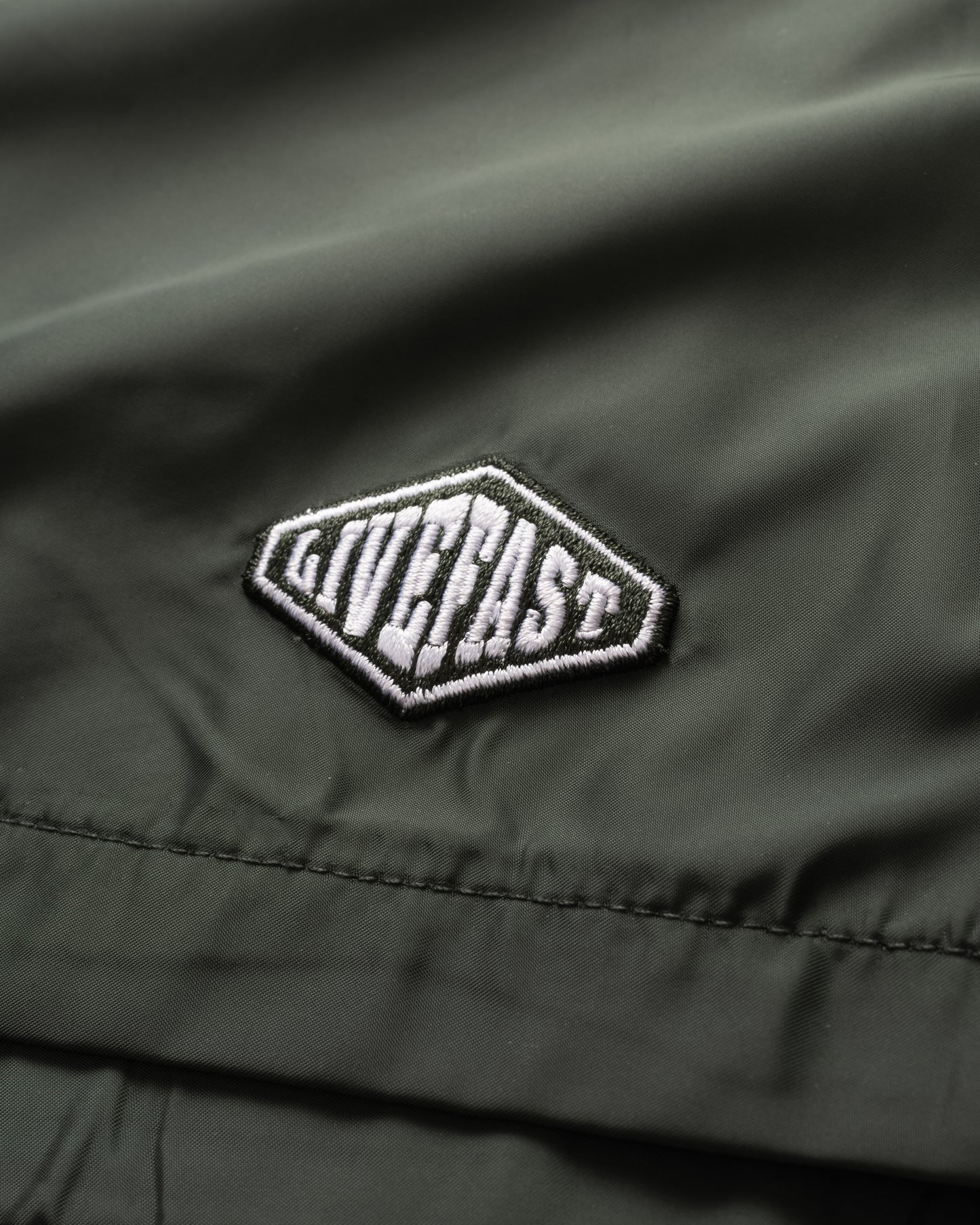 A close-up of olive green Basic Satin Shorts by LIVE FAST DIE YOUNG in polyester fabric features a white and black embroidered logo patch and a comfortable elastic waistband.