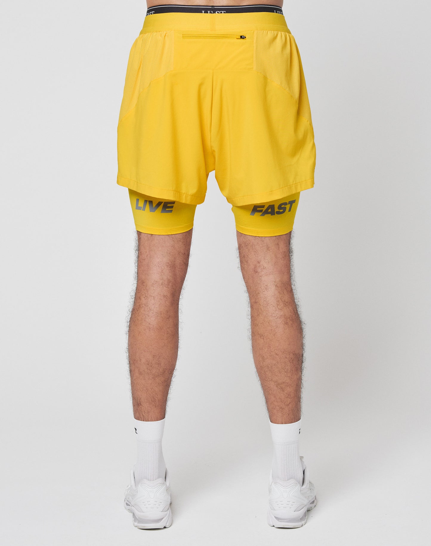 A person stands facing away, wearing LIVE FAST DIE YOUNG’s Live Fast Performance Running Shorts with integrated tights and branding on the legs, paired with white socks and sneakers against a plain background.
