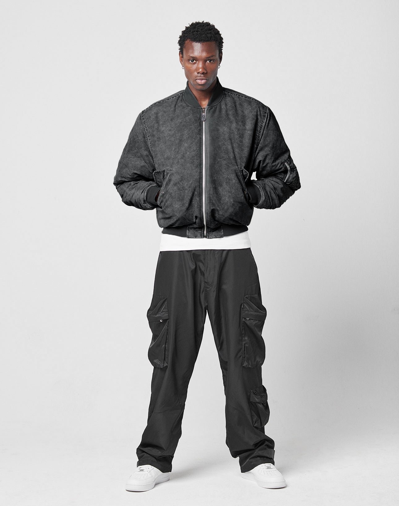 A person stands against a plain light gray background wearing the LIVE FAST DIE YOUNG Washed Bomber Jacket, black cargo pants, a white shirt, and white sneakers. Their hands are in their jacket pockets.