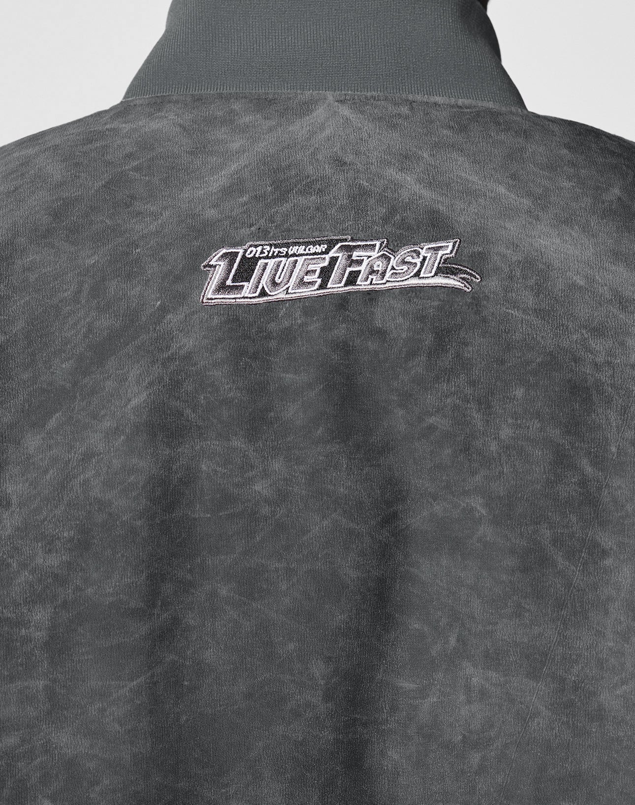 A close-up of the back of a gray Washed Bomber Jacket by LIVE FAST DIE YOUNG, featuring textured fabric and white and pink embroidery reading Live Fast with smaller text above that says 0137 COLLDN.