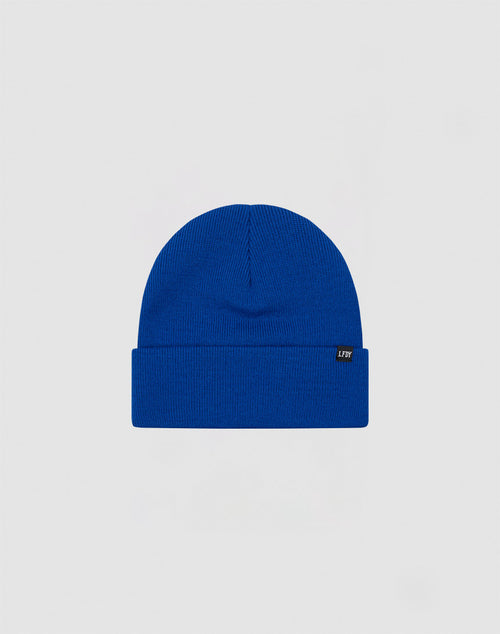 The LIVE FAST DIE YOUNG Basic Beanie is a plain royal blue polyacryl hat with a folded brim and a small black side flag label with white text, shown on a light gray background.