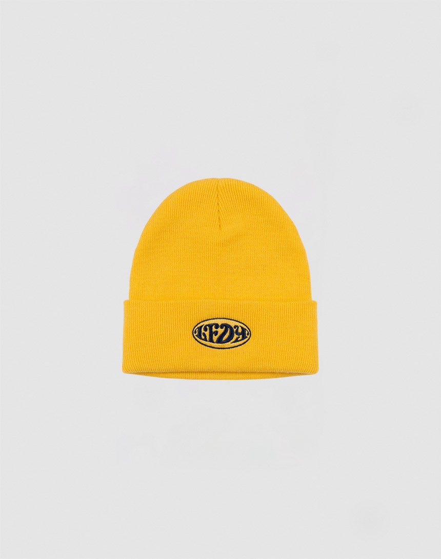 The LFDY Oval Beanie by LFDY is a yellow polyacryl knit hat with a folded brim and a small black oval patch with white text on the front, shown against a plain light gray background.