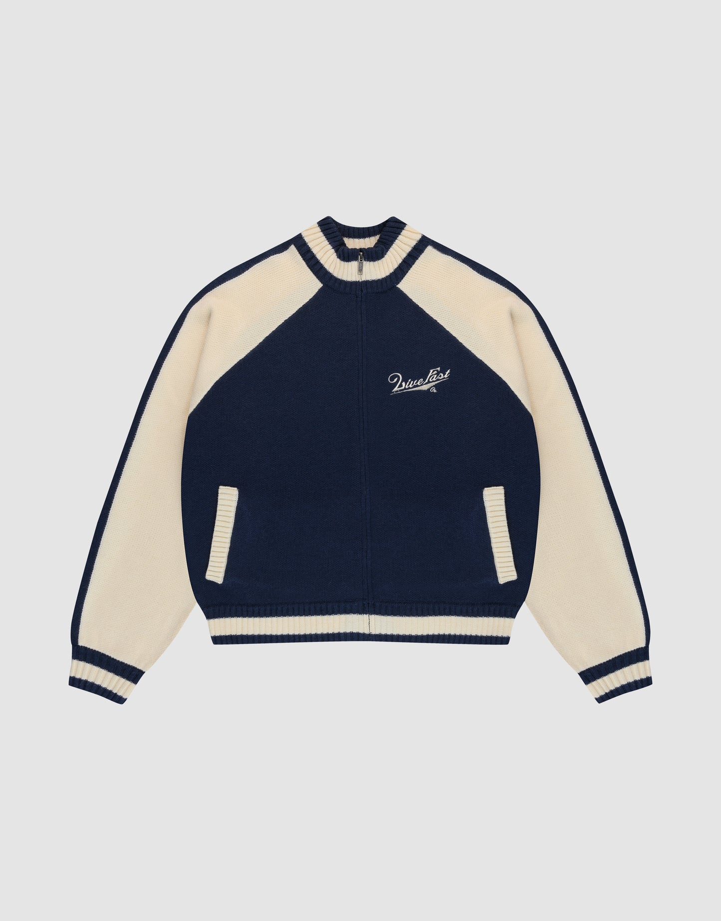 The LIVE FAST DIE YOUNG Boxy Knit Zip Jacket features a navy and cream varsity style with cream raglan sleeves, striped cuffs and collar, two front pockets, and Paradise embroidery on the left chest. Set against a plain light gray background.