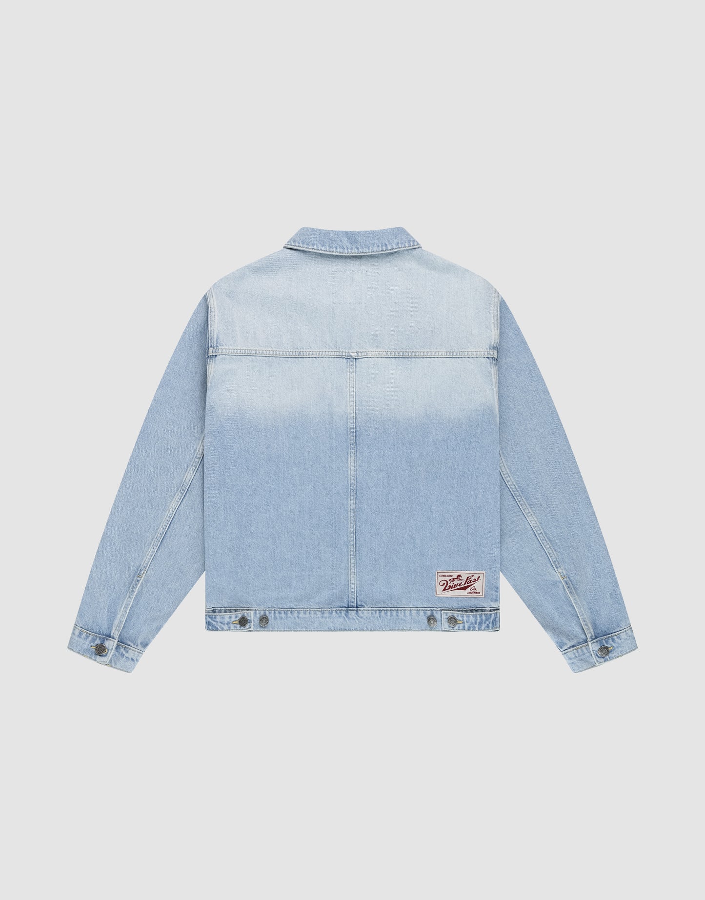 The LFDY Trucker Jacket by LIVE FAST DIE YOUNG is a light blue denim jacket shown from the back, featuring a small white and red patch near the bottom right hem, classic collar, long sleeves, cotton fabric, and adjustable button tabs with subtle rivet details.