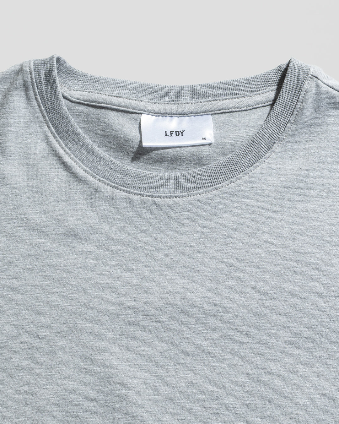 A close-up of the LFDY Slim Tee by LIVE FAST DIE YOUNG in light grey, featuring a white neck label with brand and size M, plus a signature Flag-Label. The slim fit T-shirt is shown laid flat on a light background.