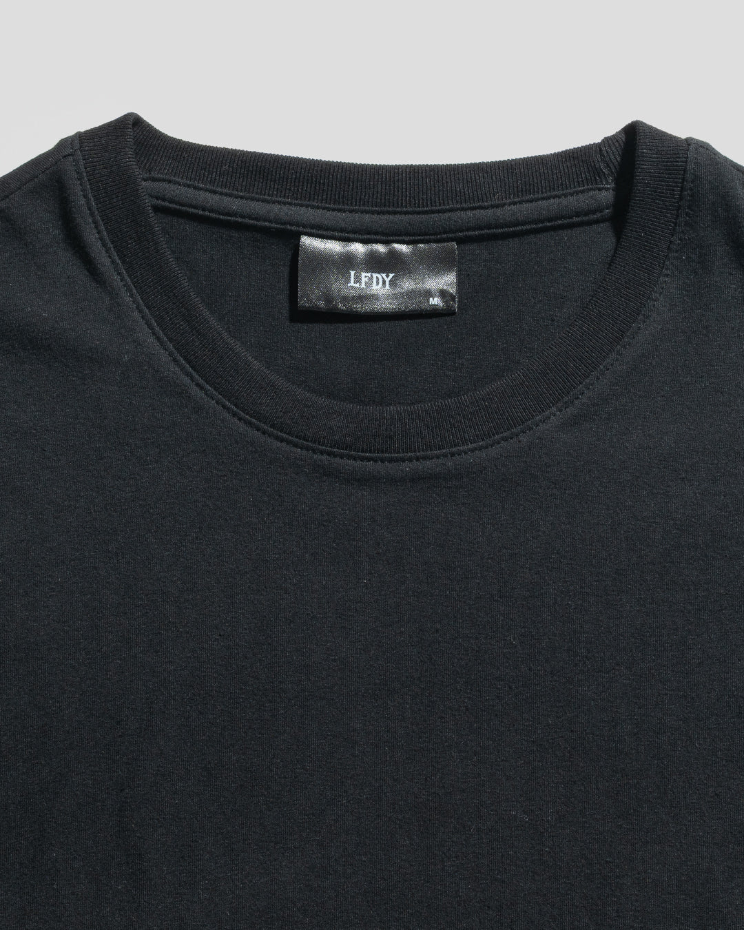 Close-up of a plain black LFDY Slim Tee by LIVE FAST DIE YOUNG, lying flat to reveal the upper chest and neckline. A black label with "LFDY" text is visible inside the collar. The shirt is crafted from soft cotton.