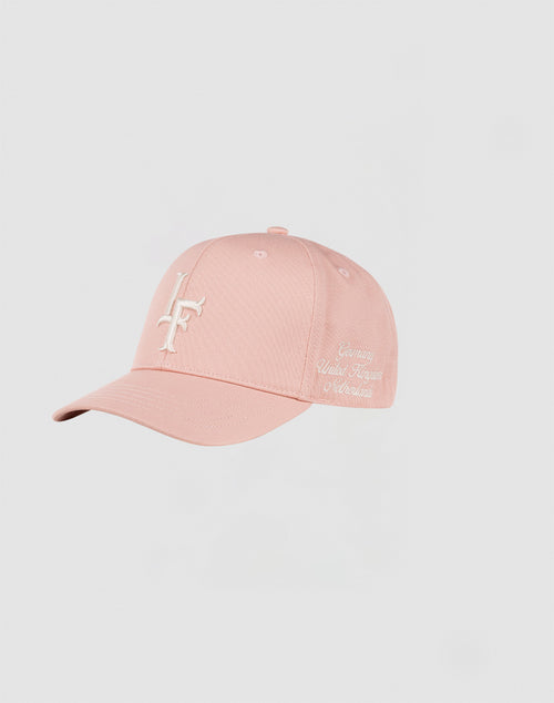The LF Snapback by LIVE FAST DIE YOUNG is a light pink cotton cap featuring a white embroidered LF logo on the front and Genuine Reality Forged Of Motivation in cursive on the side, with a snap closure, shown on a light gray background.