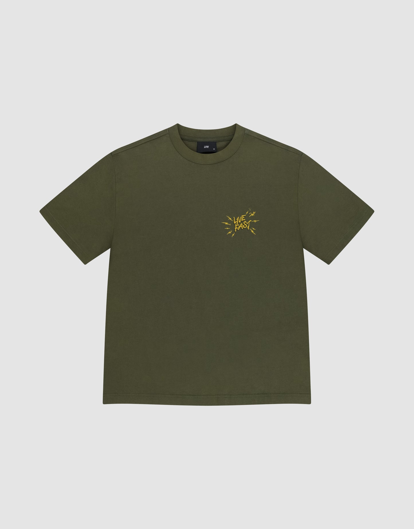 Das LFDY Against Us Tee von LIVE FAST DIE YOUNG ist ein olivgrünes Oversize-T-Shirt aus 100% Baumwolle mit gelbem Frontprint und "LIVE FAST" Schriftzug auf der linken Brust, präsentiert vor hellem Hintergrund.