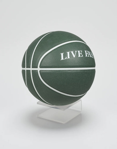 The Live Fast Basketball by LIVE FAST DIE YOUNG is dark green with white lines, made from hygroskopisches PU and has an extra griffige Oberfläche. It features LIVE FAST branding and is shown on a minimalist clear stand against a white background.