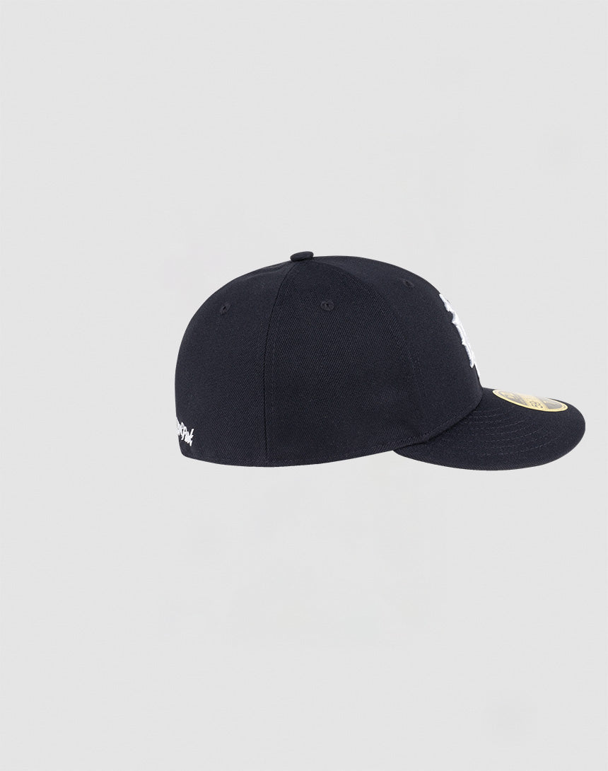 The LFDY x NEW ERA 59FIFTY Low Profile fitted cap by LIVE FAST DIE YOUNG is black, viewed from the side, with a gold sticker on its flat brim and white side embroidery, set against a plain light gray background.