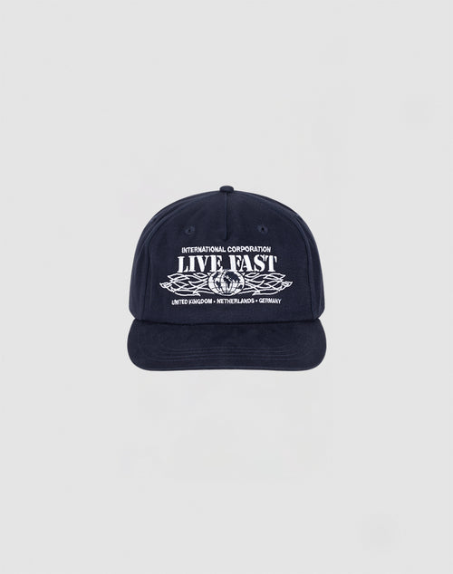 The Live Fast Corporation Cap by LIVE FAST DIE YOUNG is a navy unstructured 5-panel hat with INTERNATIONAL CORPORATION LIVE FAST in white, leaf graphic, multi-logo accents, and a flag label listing UNITED KINGDOM, NETHERLANDS, and GERMANY.